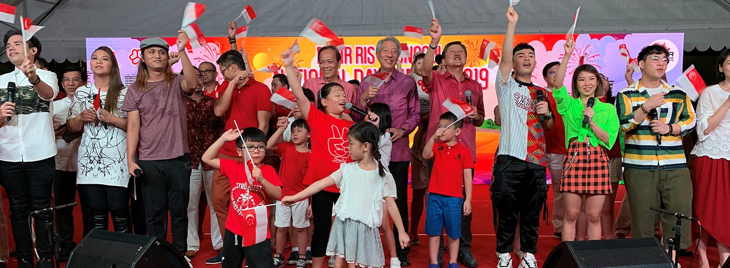 Group of people on stage waving Singapore flags in front of "Our Singapore" backdrop.